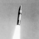 A black-and-white photo of a missile blasting upwards through the air with flames propelling it toward its target