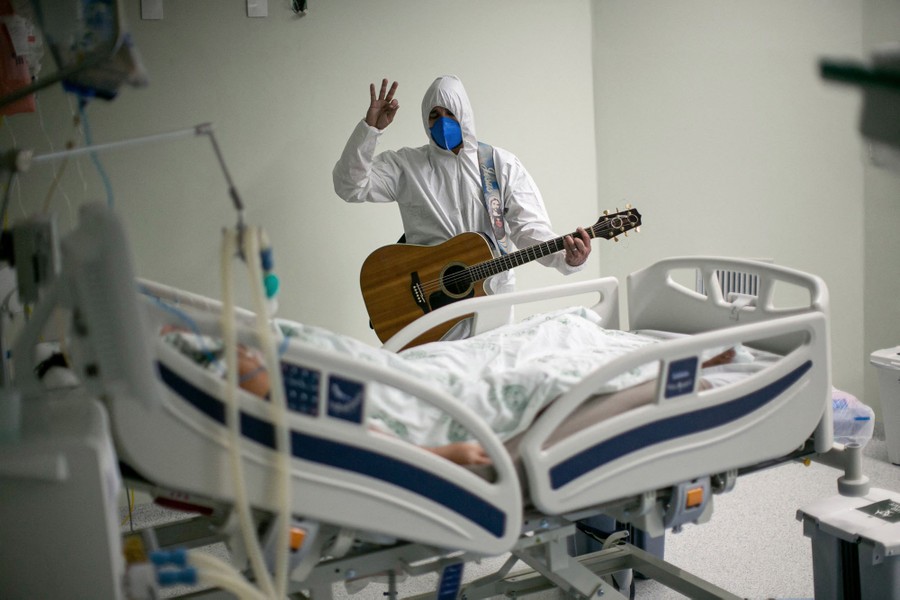 A man wearing protective equipment performs with a guitar for a patient.