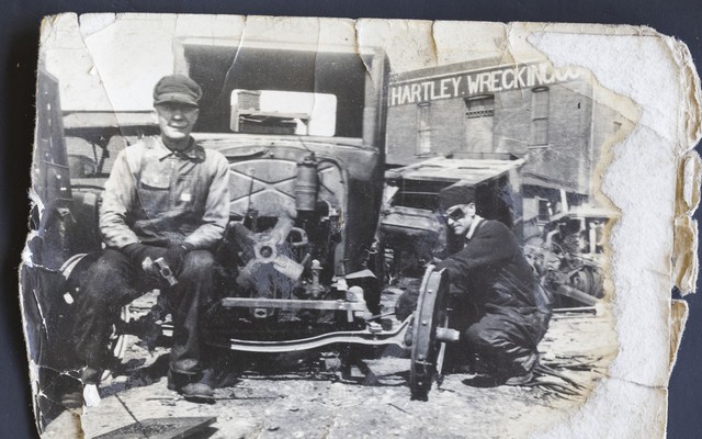 A black-and-white photo of a man sitting on an old-fashion car in the 1930s or 1940s