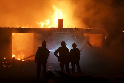 Firefighters stand in front of a burning house.