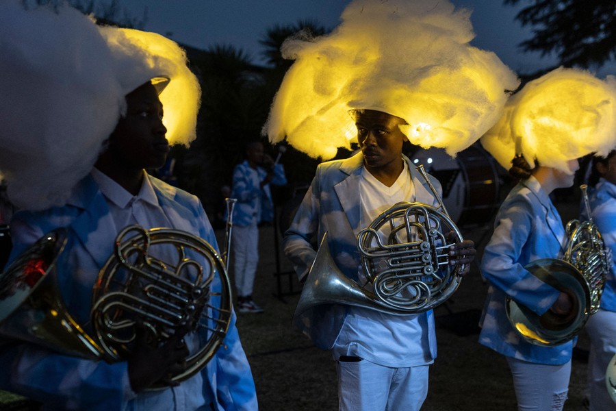 Performers wearing illuminated cloud-shaped headwear carry French horns.