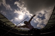 An athlete stretches and leans back while preparing to throw a javelin inside a stadium, silhouetted against a cloudy sky.