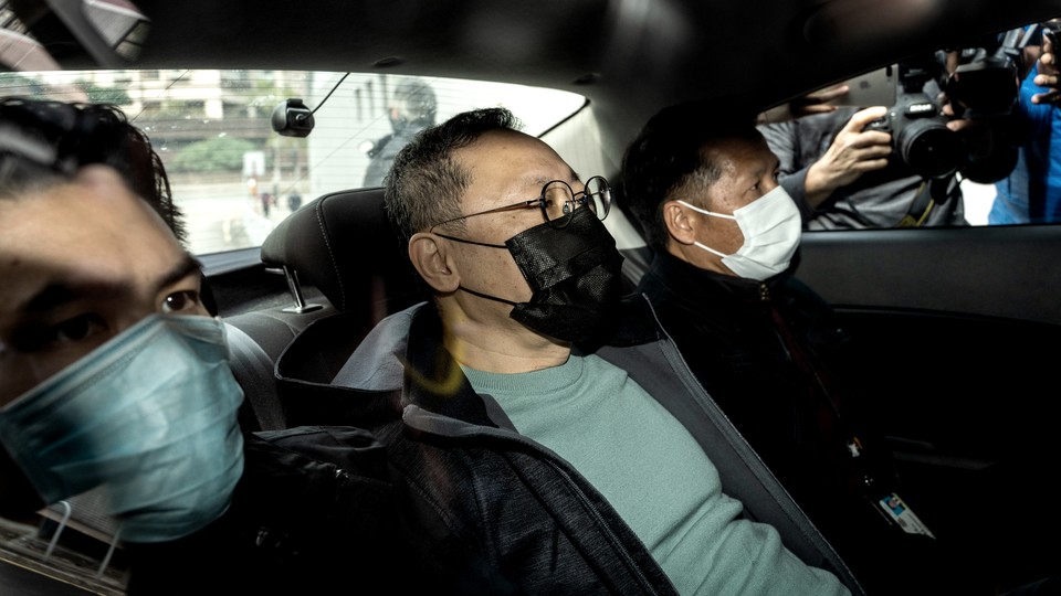 Three men sit in the backseat of a car with masks on as photographers stand outside.