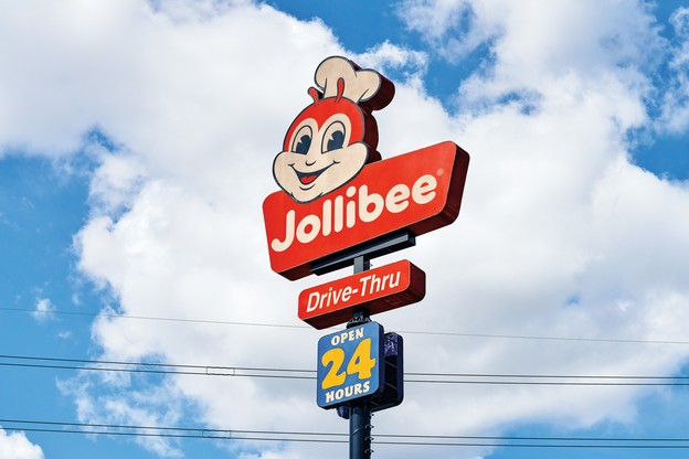 photo of roadside red Jollibee sign with smiling bee, Drive-Thru, and open 24 hours on it, with bright blue sky and clouds in background
