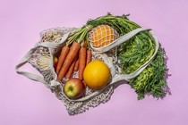 A woven bag containing carrots, apples, two oranges, and some greens, against a lavender background