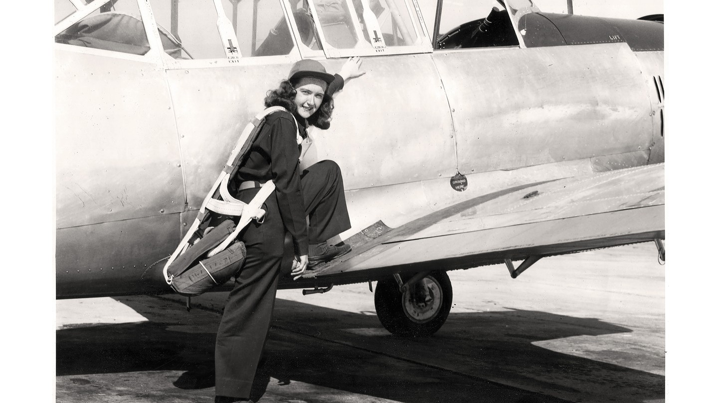 black-and-white archival photo of woman in flight gear and cap, smiling at camera, standing with one foot on ground and one foot on the wing of the plane she piloted, about to climb into cockpit