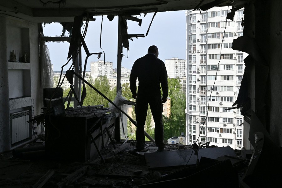 A man stands in a bomb-damaged apartment, looking out through the missing outer wall.