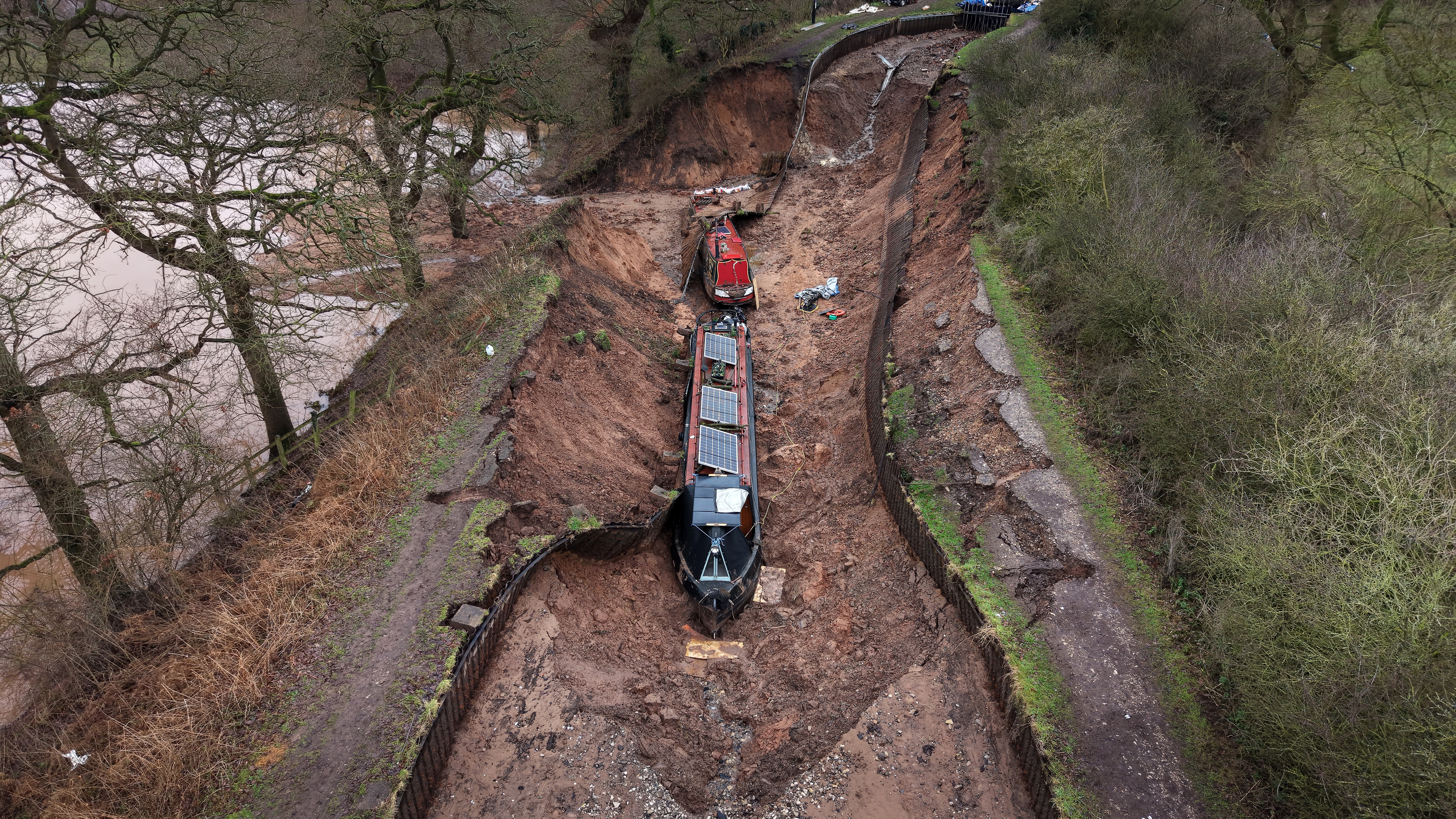 Two long canal boats sit on the bottom of an emptied section of canal, near a breach in an embankment.