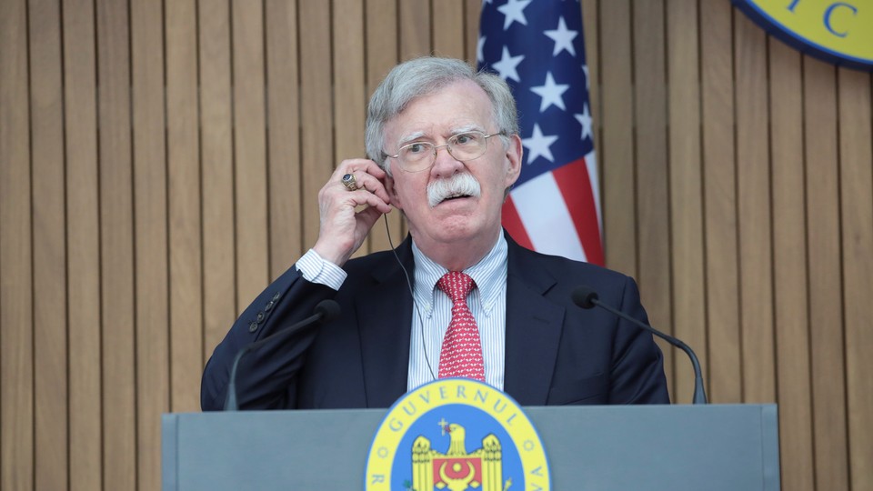 John Bolton stands in front of a podium and an American flag.
