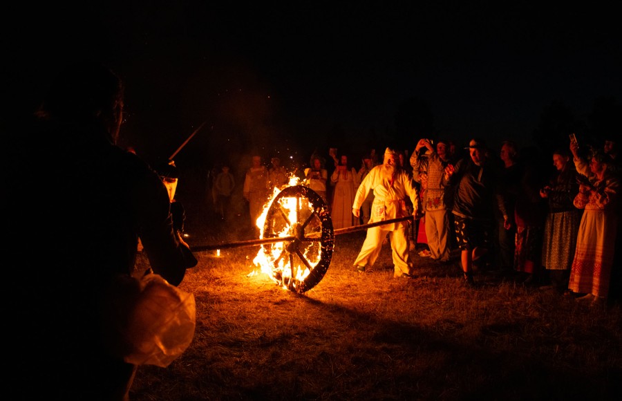 A crowd looks on as two people in traditional costumes use a long pole to roll a burning wheel across a grassy area at night.