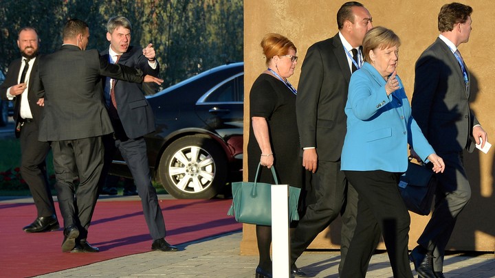 German Chancellor Angela Merkel arrives in Marrakech for the signing of a United Nations migration compact as man gestures angrily in the background.