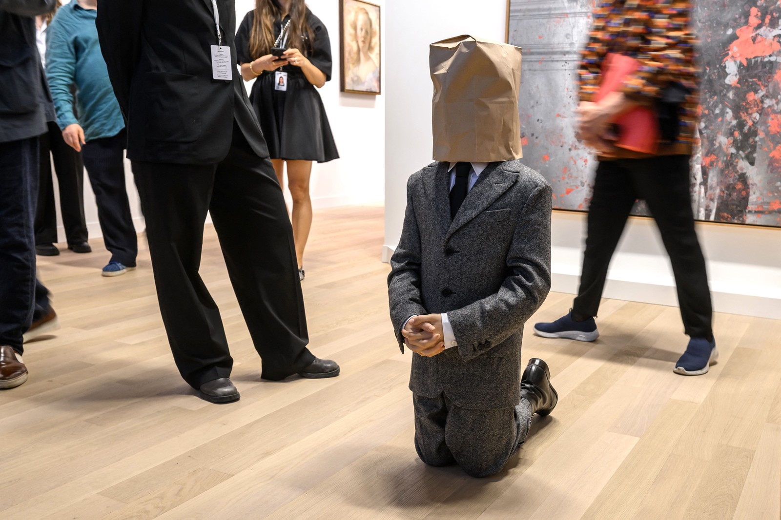 In an art gallery, people walk about and look at artwork, including a small-scale statue of a man wearing a suit that is depicted as kneeling while wearing a paper bag over its head.