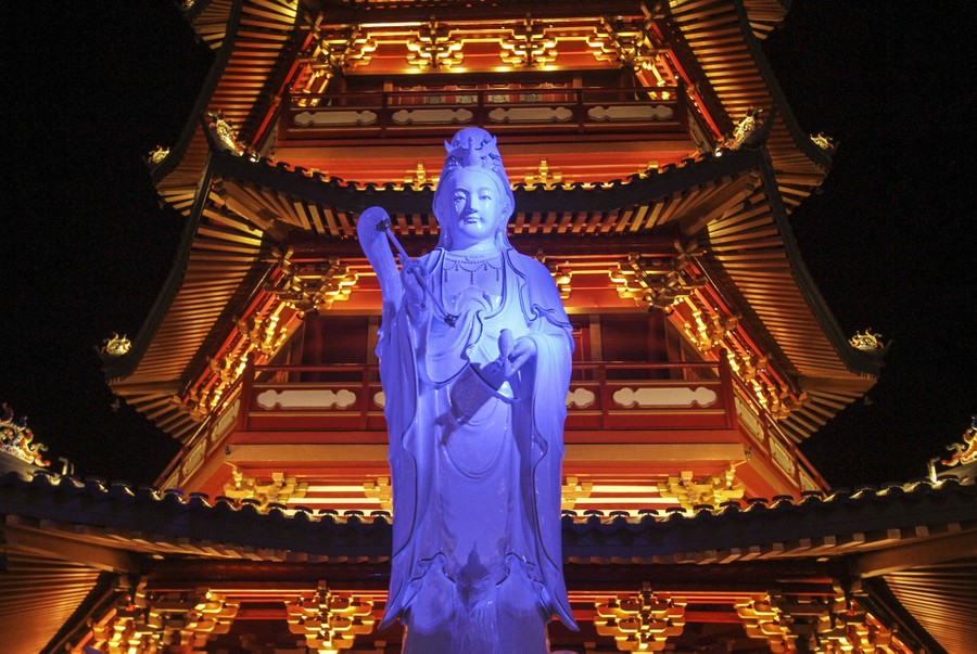 An illuminated statue stands in front of an ornate pagoda-style building at night.