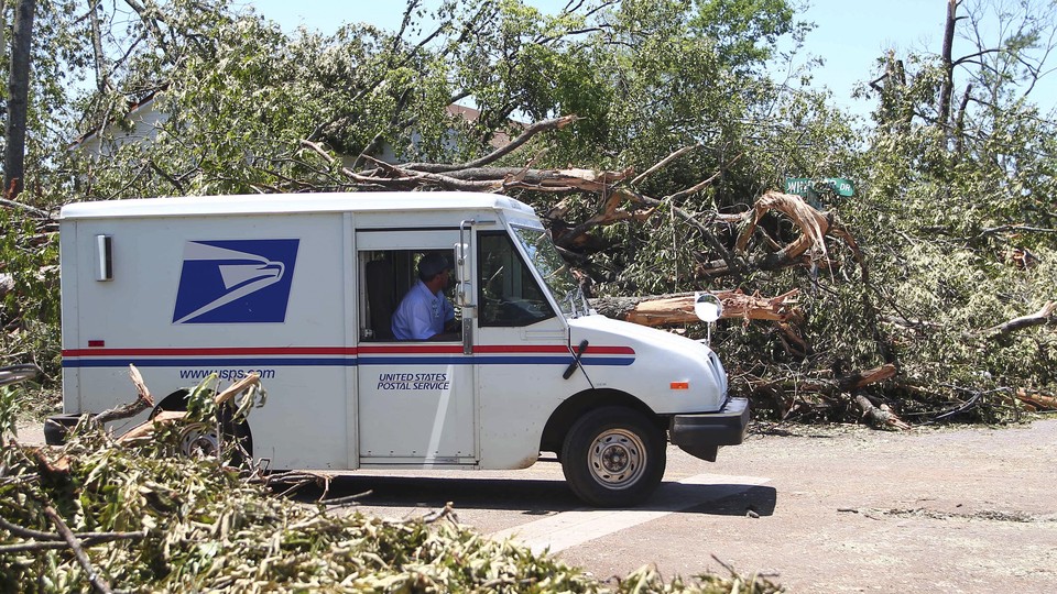 A U.S. Postal Service truck drives down a road lined by downed trees.