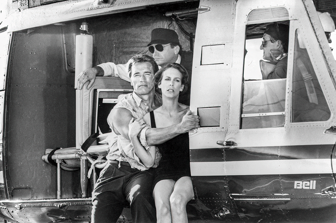 black-and-white photo still from movie set of Arnold with arm around Jamie Lee Curtis sitting in doorway of ambulance 