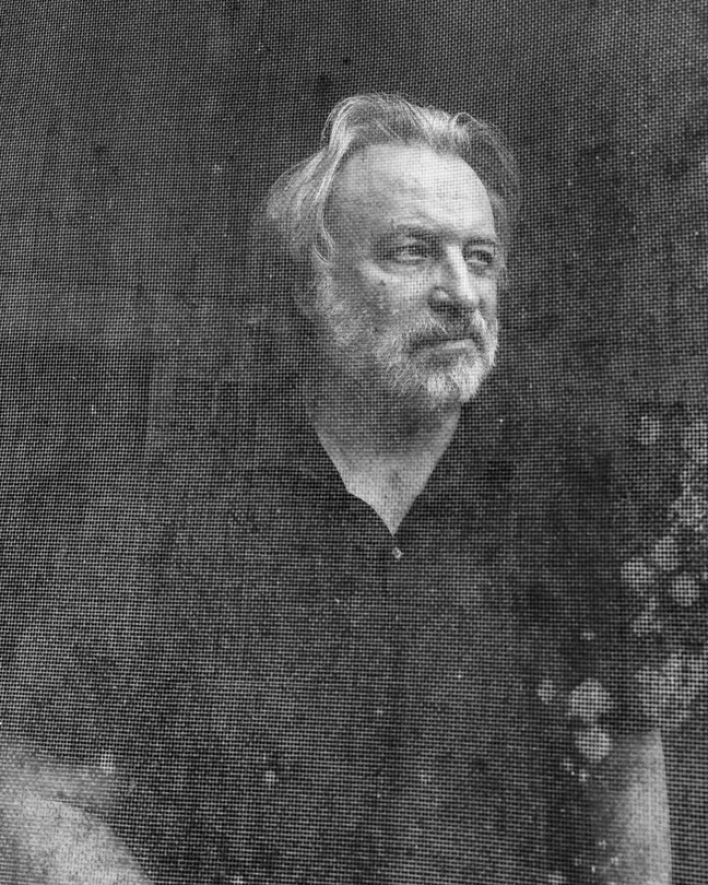 Black and white photo of a man in a black short sleeved shirt with a gray beard and swooping hair.