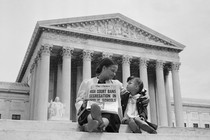 A black-and-white photograph of a woman and child sitting on the steps of the Supreme Court, holding up a newspaper with the front page headline, 'HIGH COURT BANS SEGREGATION IN PUBLIC SCHOOLS'