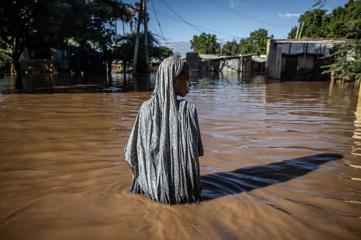 A woman stands waist-deep in muddy floodwater, surrounded by partially submerged houses and trees.