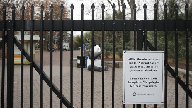 A white sign on a black gate says that the National Zoo is closed due to a government shutdown. In the background what appears to be an inflatable panda bear sits on the path in the empty zoo. 