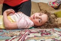 Girl with curly hair lying on colorful carpet with head turned toward the camera, with someone kneeling next to her and couch in background