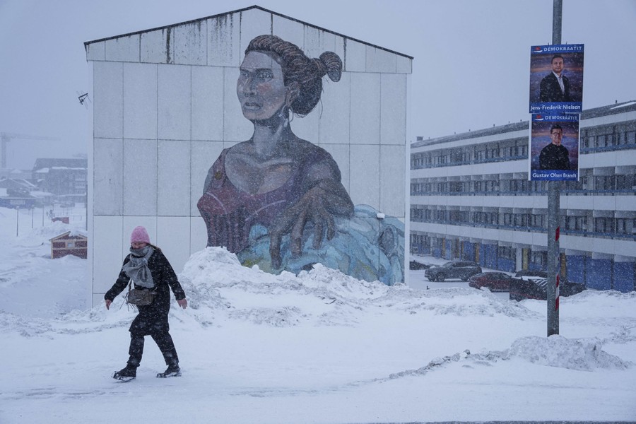 A woman walks on a snow-covered street past a mural painted on a building and two political posters on a post.