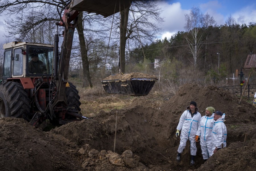 Three people wearing protective gear stand inside a pit as a tractor lifts a casket from the ground.