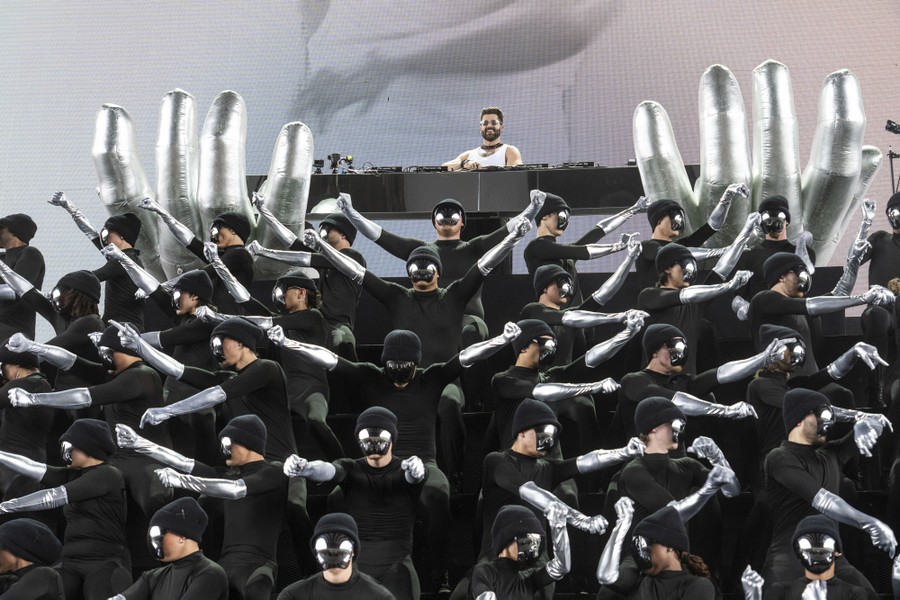 A performer stands on stage, behind a large group of dancers who perform choreographed moves, wearing long silver gloves.