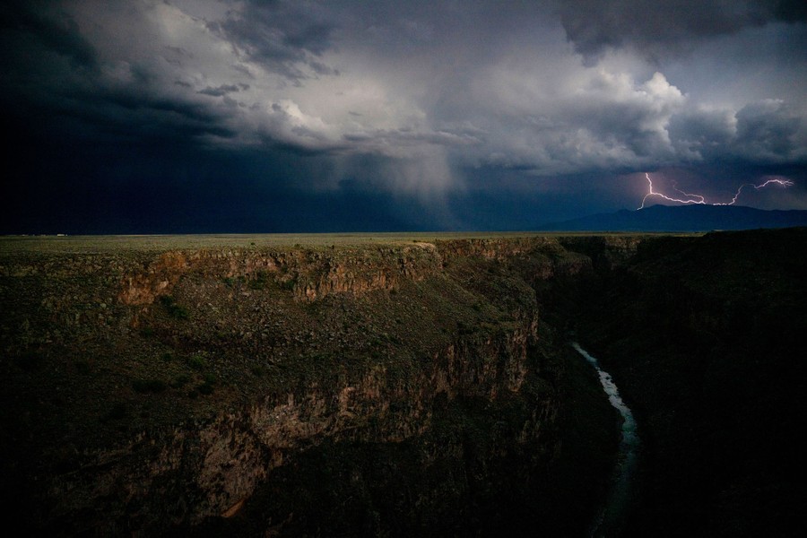 Stormy clouds and lightning appear above a river canyon.
