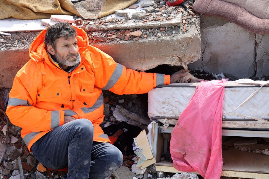 A man sits beside rubble, his left arm reaching out and holding the hand of his daughter, who lies dead, trapped beneath the debris.