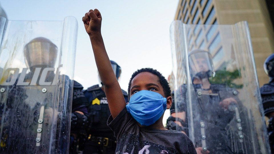 A young black boy wearing a mask raises his fist in front of a line of police officers using riot shields