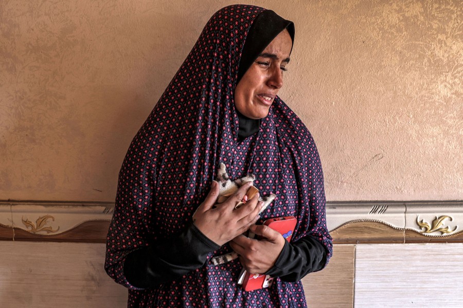 A woman stands against an interior wall of a house, emotional, holding small personal items.