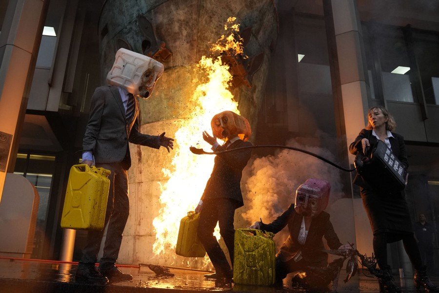 Four people, including three wearing fuel-can masks, protest in front of a fire outside a building.