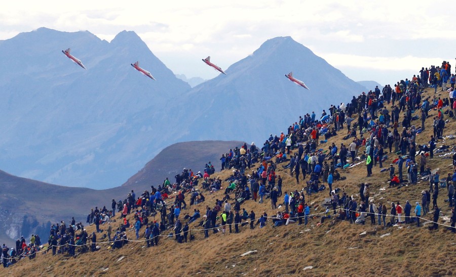 Four fighter jets fly among mountains, close to a gathered audience on a hillside.