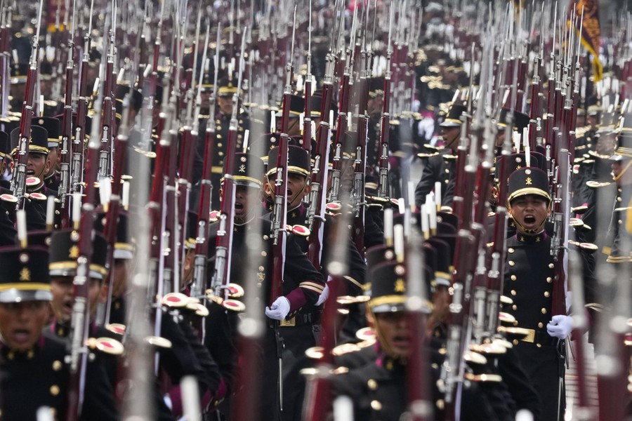Military cadets in uniform, carrying rifles with bayonets, march in a parade.