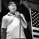a black-and-white photo of a man speaking into a microphone next to an American flag