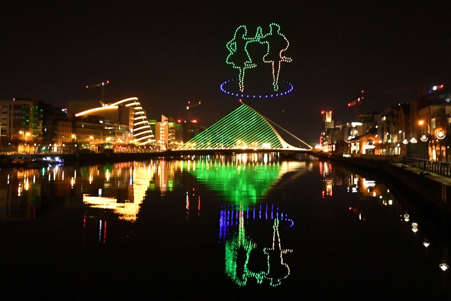 The illuminated figures of two dancers appear in the sky above the lit-up buildings of Dublin, at night.