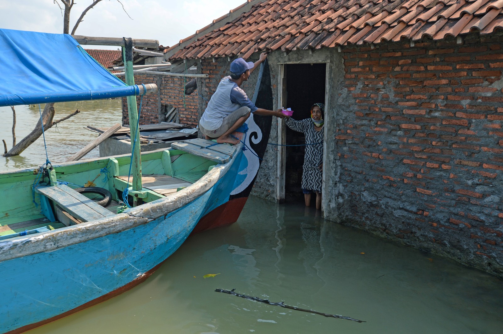 A woman stands in ankle-deep water in the doorway of a house, being handed a package by a man who is crouched at the front of a boat that has pulled up right to the house.