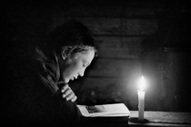 A black-and-white photo of a young boy reading by candlelight