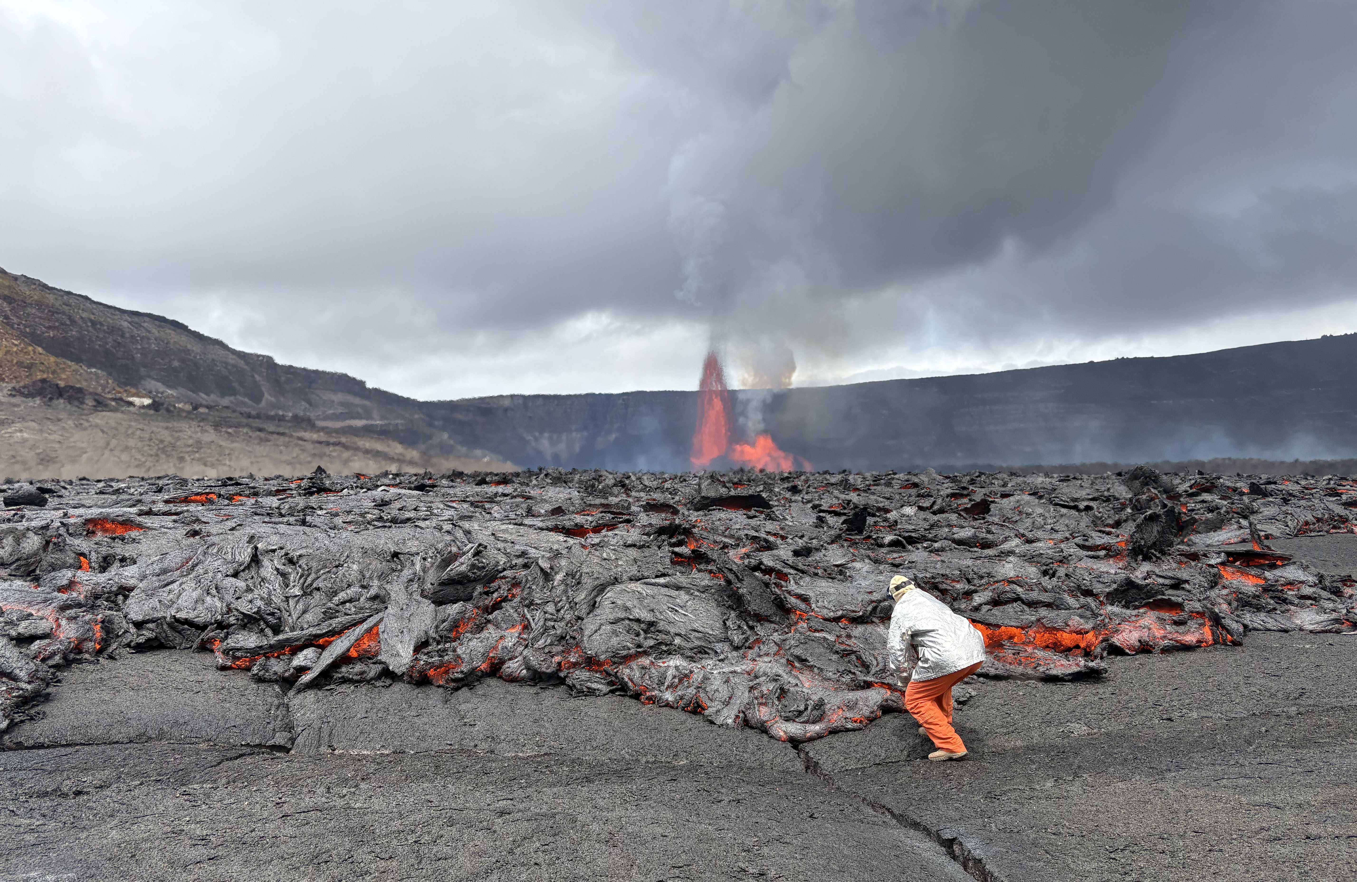 A person in protective gear leans close to an active lava flow, reaching out to gather a sample.