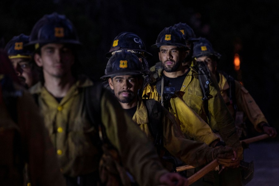 Nine or ten firefighters wearing helmets walk together under a dark sky.