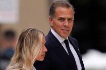 A color photograph of Hunter Biden in a dark suit and tie standing next to his wife, looking at the camera