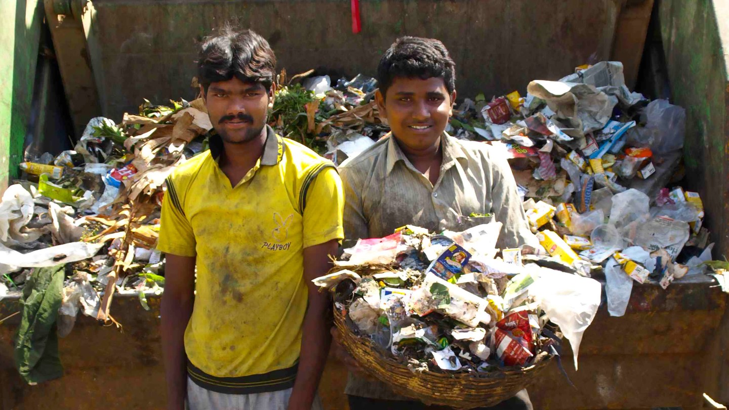 Garbage collectors in Bangalore stand in front of a garbage truck. One holds a basket of trash.