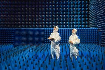 Two technicians dressed in white protective gear stand inside a test room covered in foam cones.