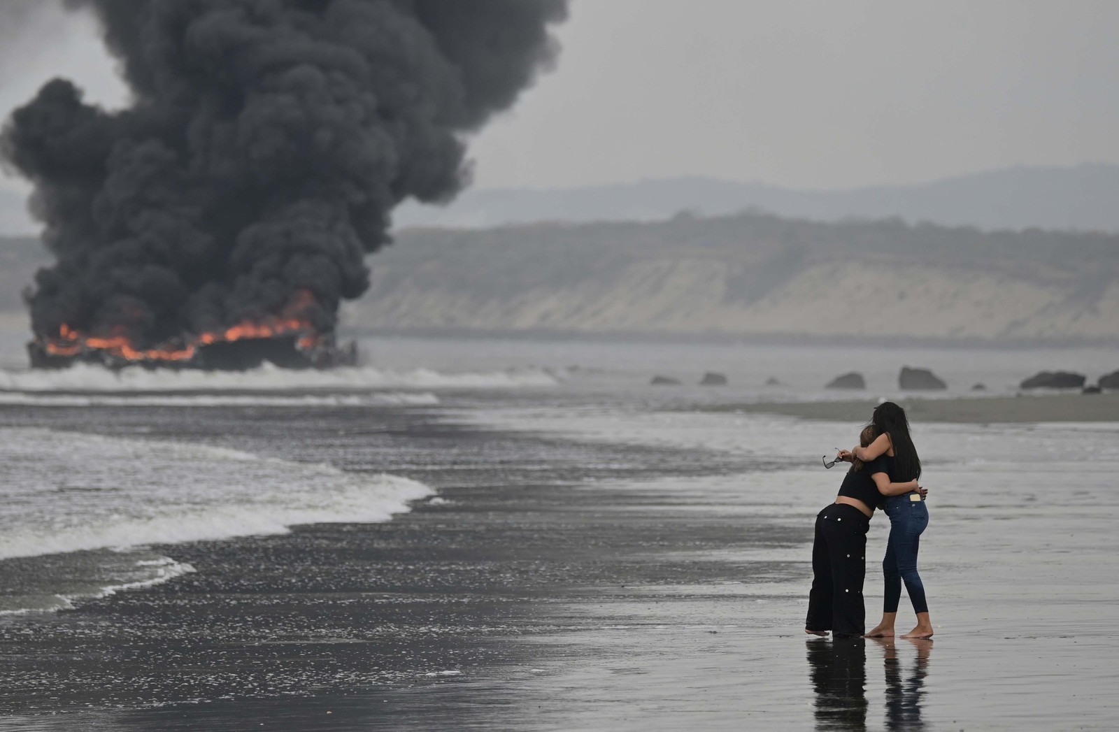 Two people embrace, standing on a beach, looking toward boats that are fully engulfed in flames in the distance.