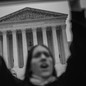Black-and-white photo of a protester, blurry in the foreground. The Supreme Court, in sharp focus, is framed by her upstretched arms.