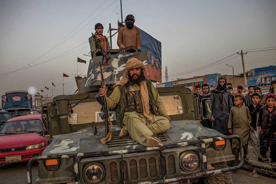 Bearded man with gun sits on hood of Humvee with 2 others standing behind with a machine gun, with other cars and crowd in background