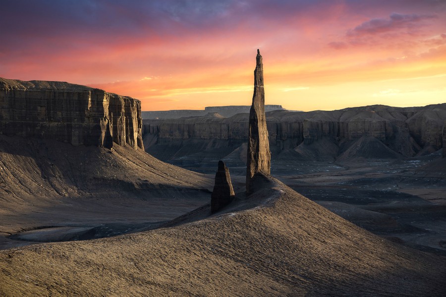 A narrow spire of stone stands in a valley amid buttes and cliffs.