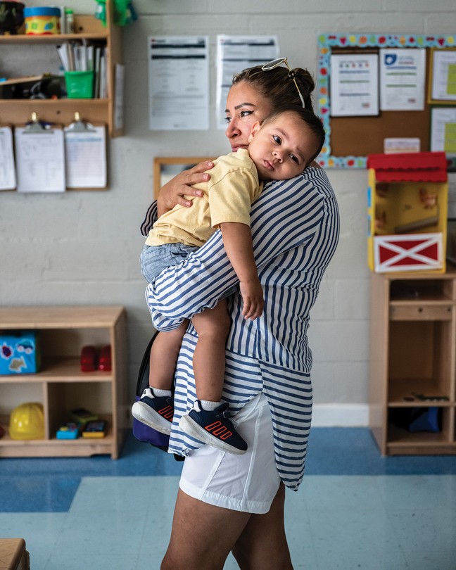 photo of woman in striped shirt and white shorts standing in children's classroom and holding toddler with his head on her shoulder