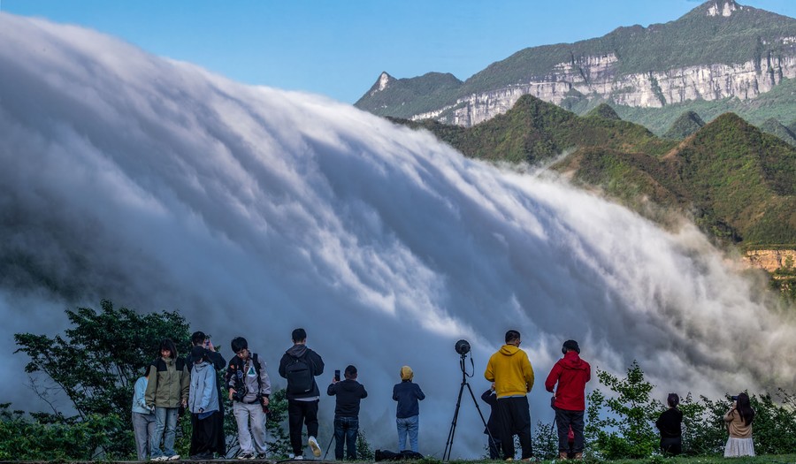 People take photographs of a low cloud formation that looks like a broad waterfall among mountains.