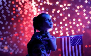 A young girl sits on the shoulders of an adult, holding an American flag, with fireworks in the background.